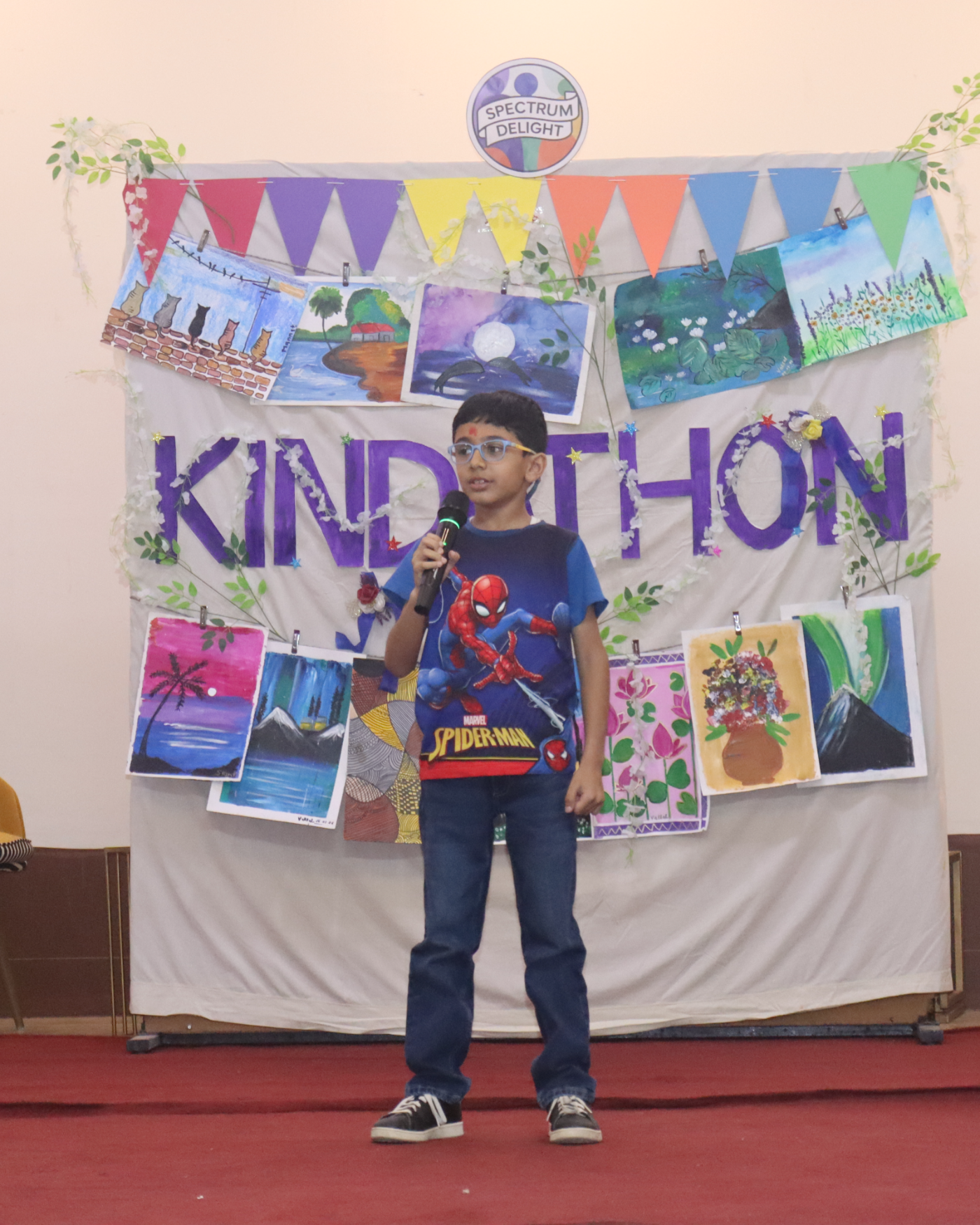 A child performing with a microphone on stage in front of a hand-painted Kindathon backdrop at a previous event